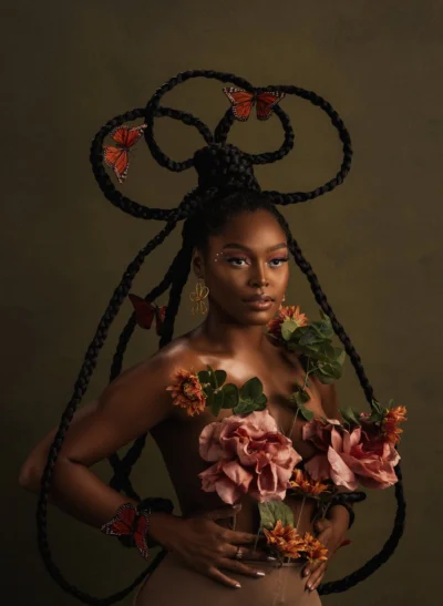A striking portrait of a woman with flowers and butterflies integrated into her hairstyle against a brown backdrop.