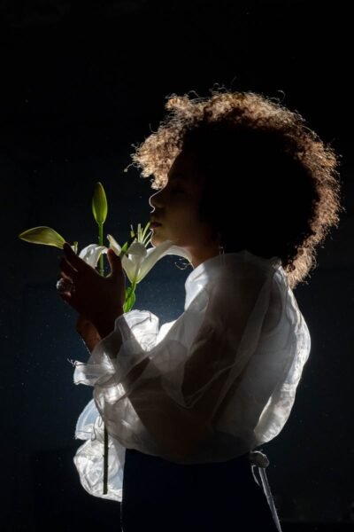 A woman in sheer clothing holds lilies in a dramatic, backlit setting, creating an artistic silhouette.